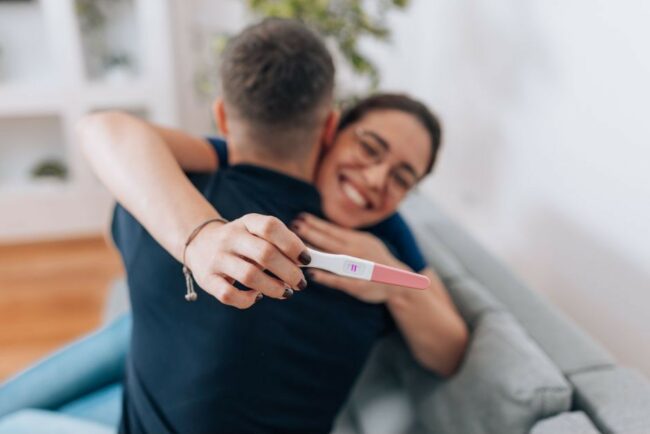 Un couple s'enlace de joie, la femme tient un test de grossesse positif